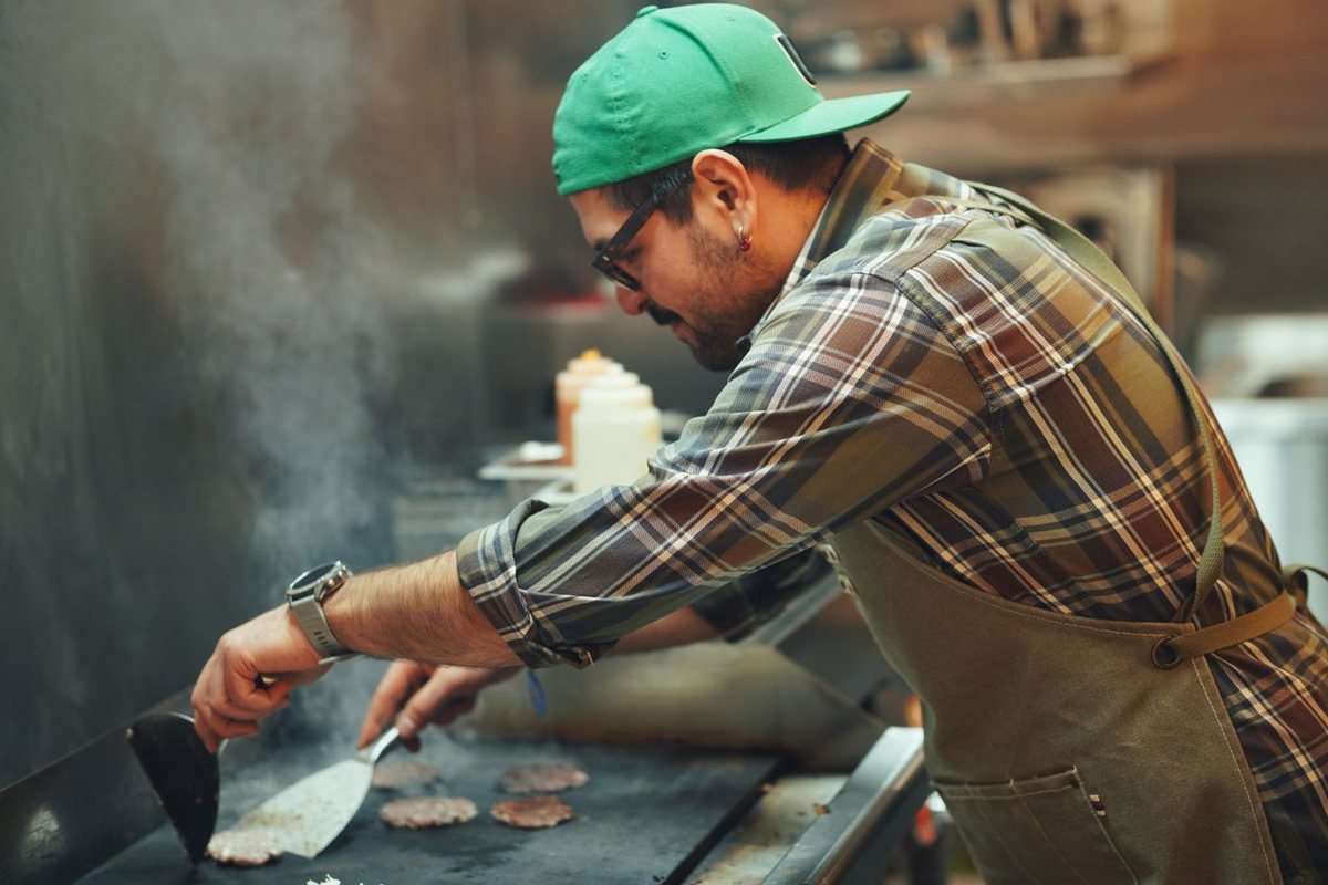 A cook in a green cap smashing burger patties on a flat-top griddle