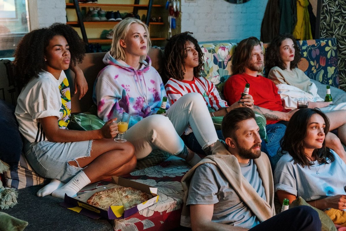 A group of friends on a couch with pizza and drinks, watching TV together